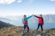 Zwei Frauen in Wanderkleidung klatschen sich die Hände ab, weil sie den Berggipfel erreicht haben. Hinter ihnen sieht man die Schweizer Alpen und einen grossen See.  Zwei Frauen in Wanderkleidung klatschen sich die Hände ab, weil sie den Berggipfel erreicht haben. Hinter ihnen sieht man die Schweizer Alpen und einen grossen See.